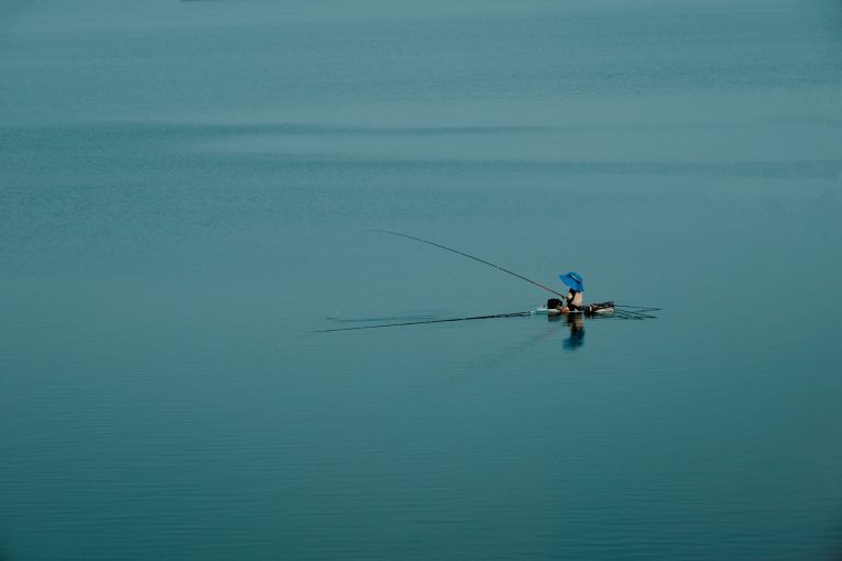A lone fisherman under a blue umbrella fishes peacefully in the middle of a vast, calm lake.