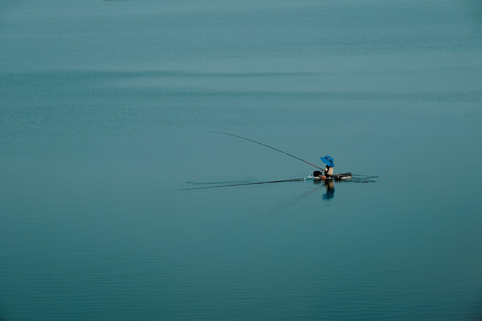 A lone fisherman under a blue umbrella fishes peacefully in the middle of a vast, calm lake.